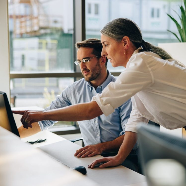 Mature woman working with young man sitting at desk and showing something on computer screen in office. Female executive pointing at desktop monitor and talking with male colleague in office.